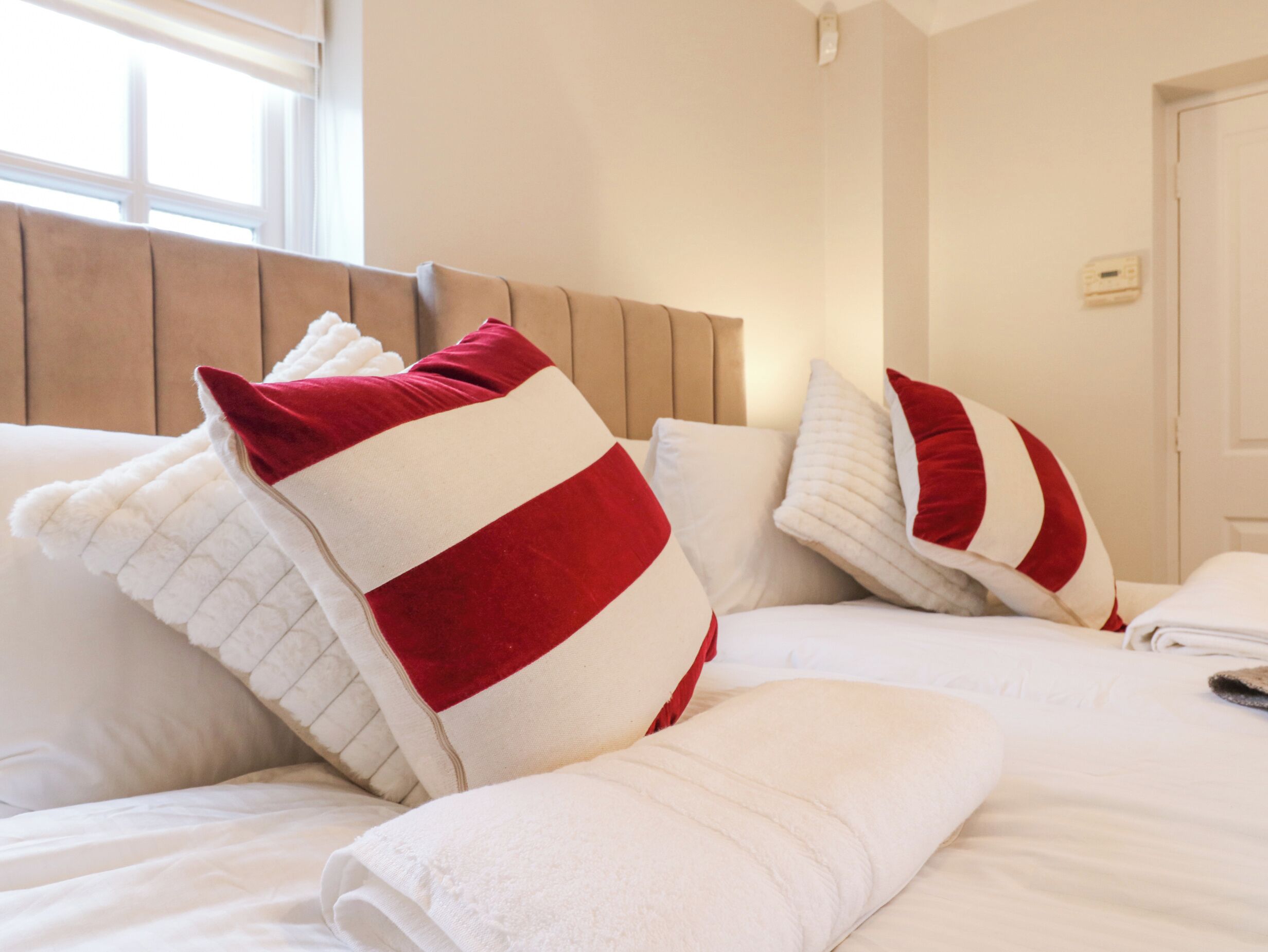 Ein ordentlich bezogenes Bett mit weiß-rot gestreiften Kissen, einem hölzernen Kopfteil und einer weißen Wand mit Fenster und Tür.