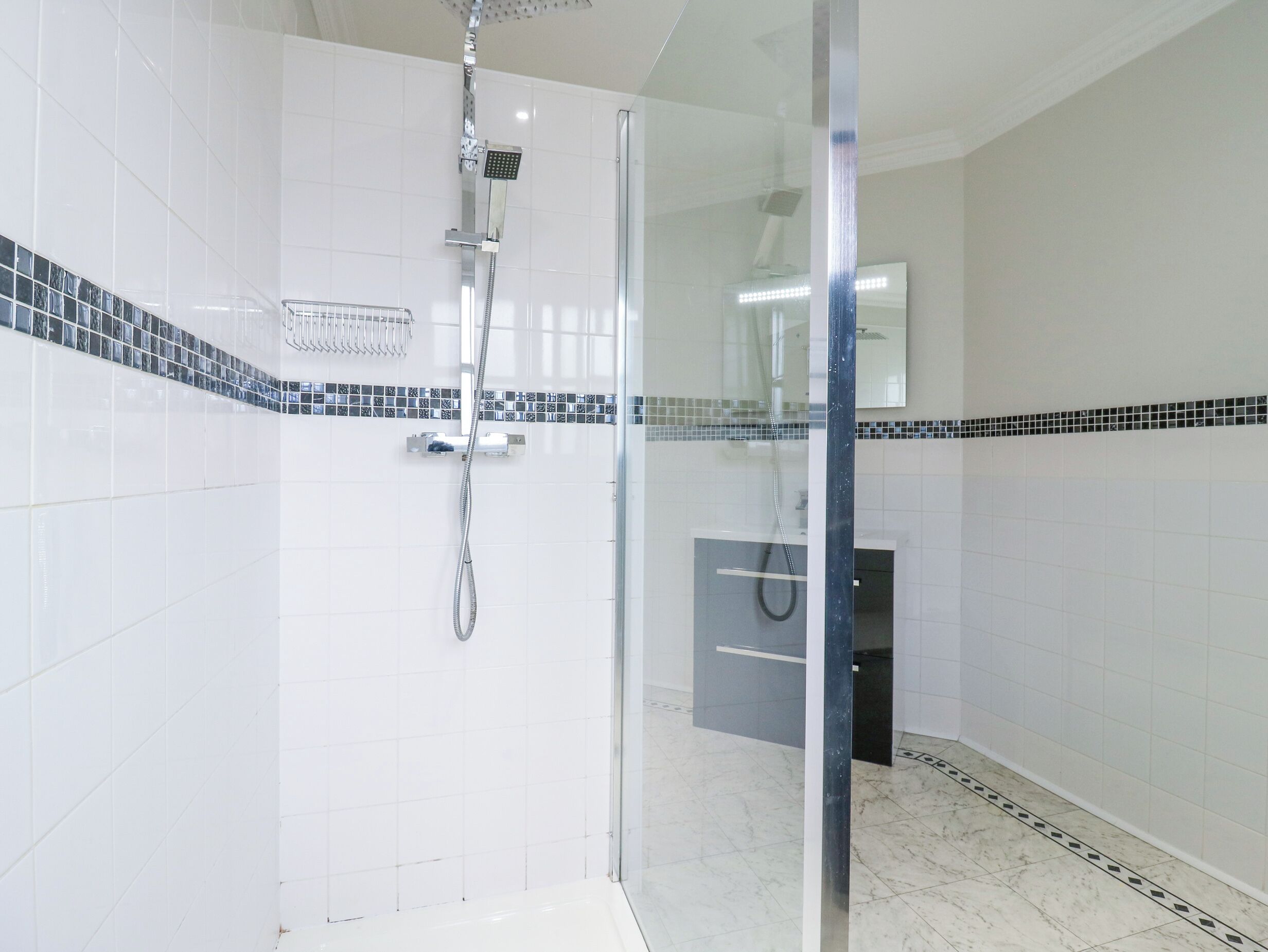 A modern bathroom with white tiles, a glass shower enclosure, a handheld showerhead, and a tiled floor.