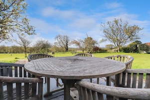 A round wooden table with four chairs, set on a patio with a view of a grassy field and trees.