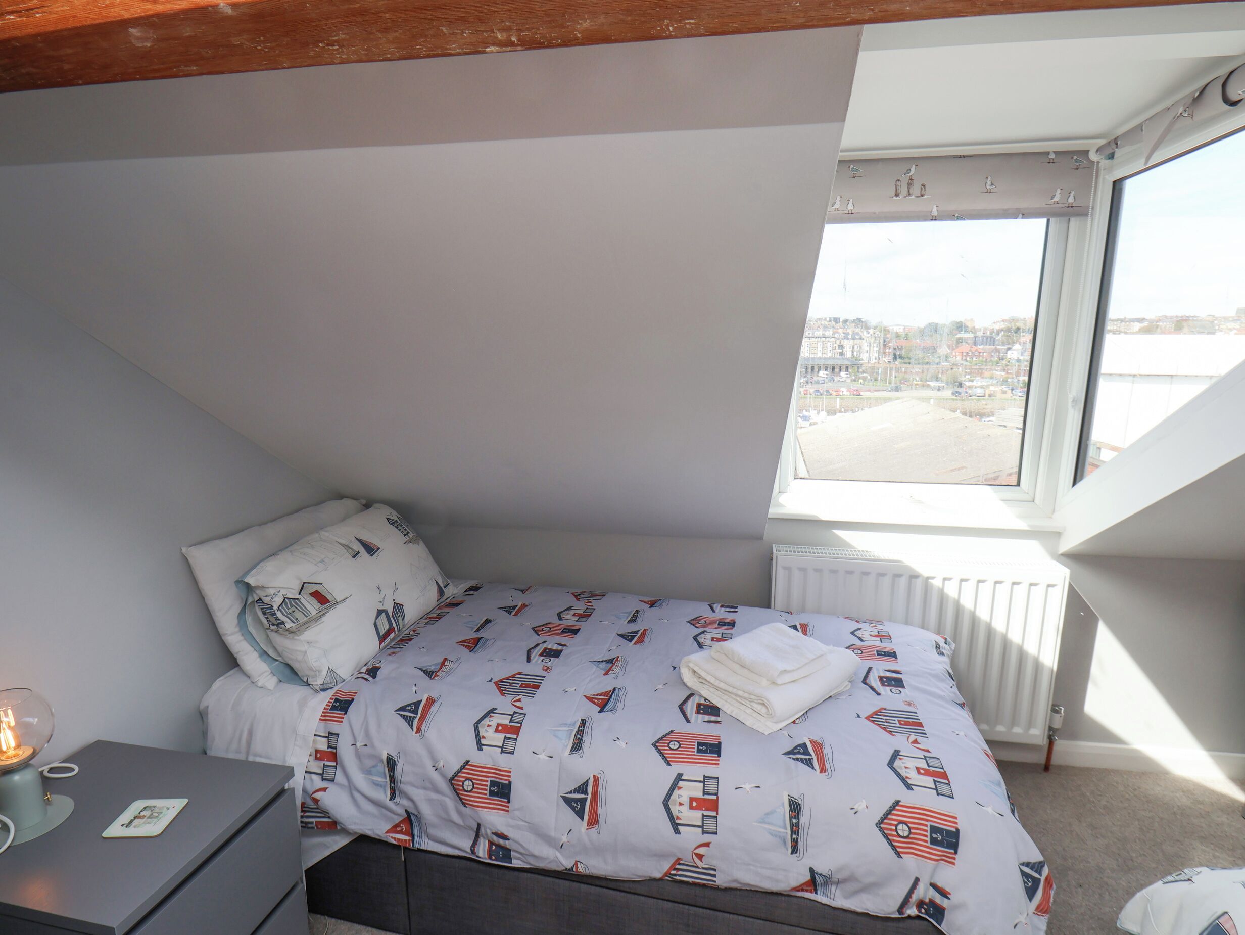 A small bedroom with a bed, a nightstand, a lamp, and a window with a city view.