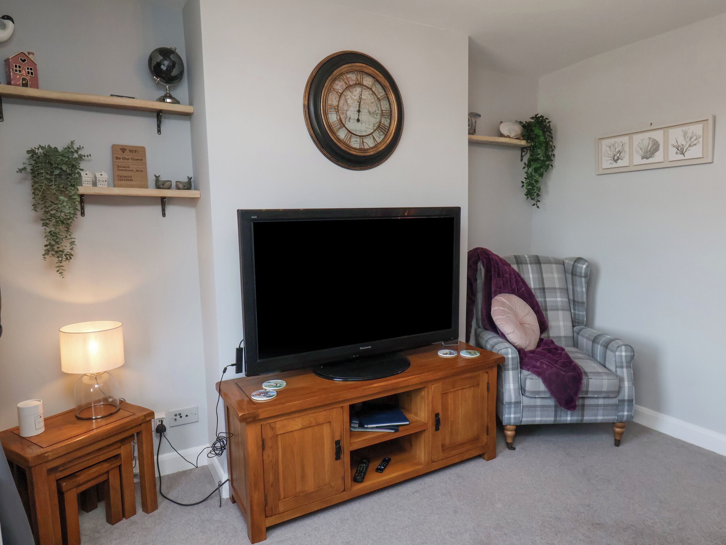 Un salon avec un téléviseur à écran plat posé sur un meuble en bois, un fauteuil à carreaux avec un plaid violet, et une horloge murale.