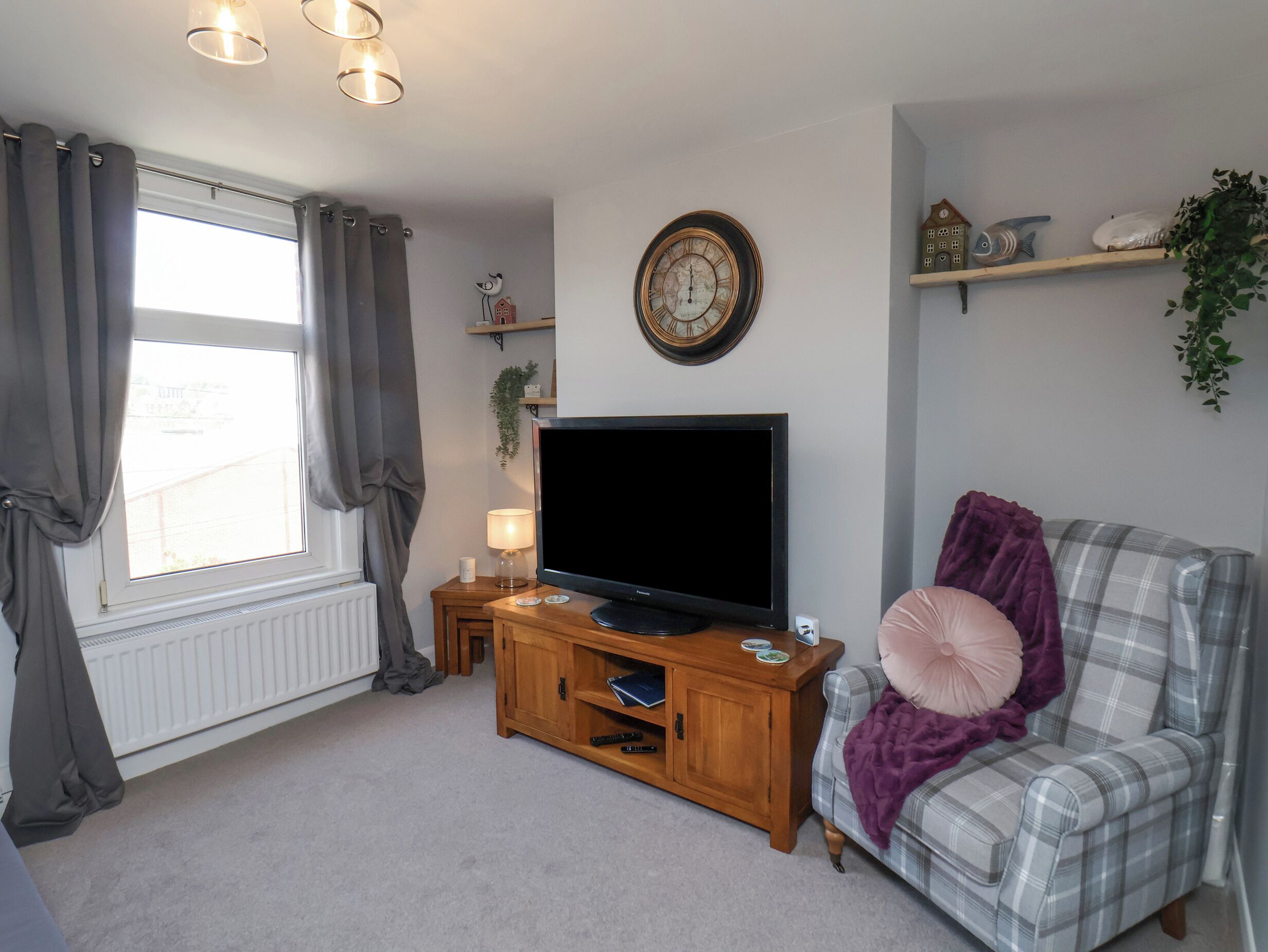 A living room with a television on a wooden stand, a plaid sofa, a clock on the wall, and a window with curtains.