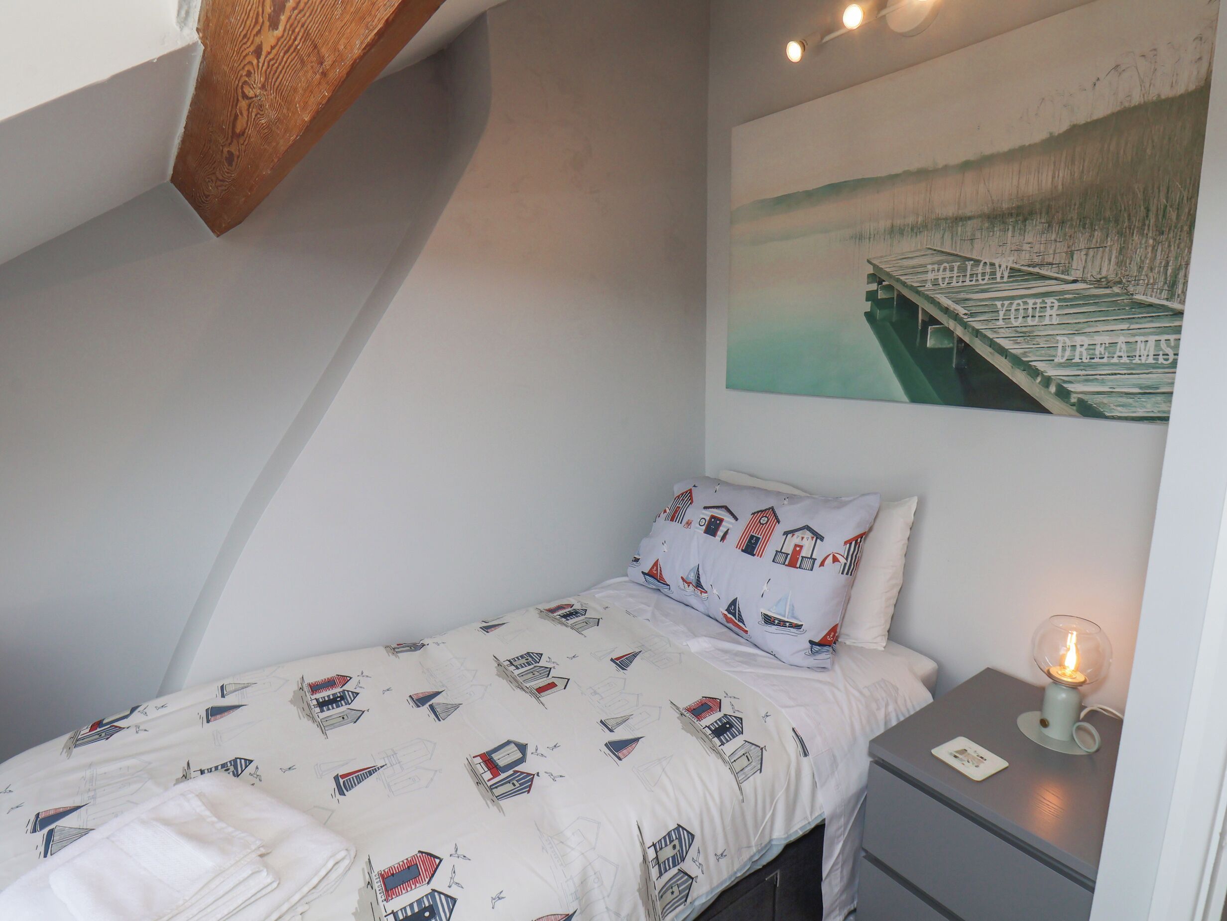 A small, neatly made bed with a patterned bedspread, a pillow with a nautical theme, a bedside table with a lamp, and a wall with a painting of a bridge over water.
