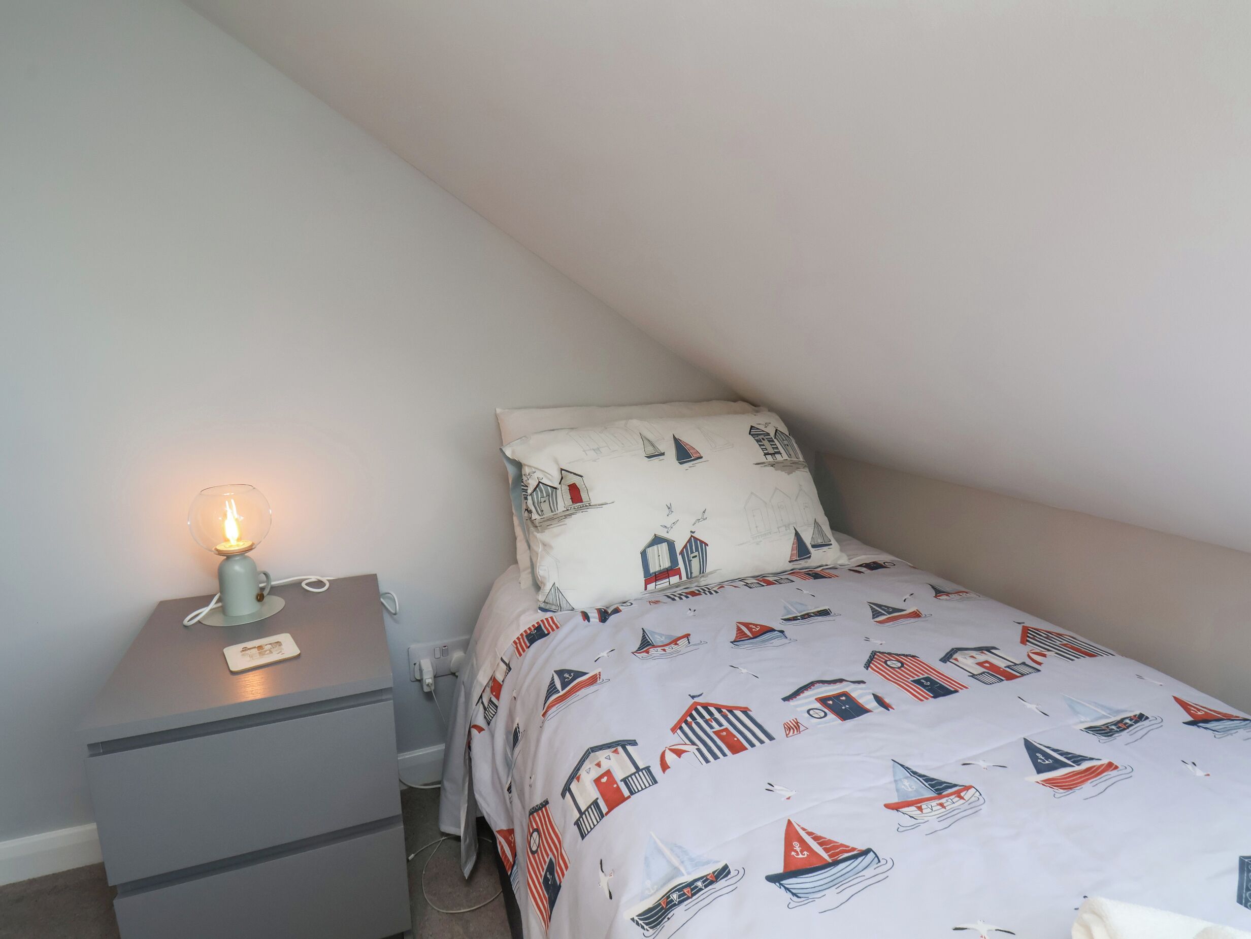 A small, neatly arranged bedroom with a single bed, a bedside table with a lamp, and a nightstand.
