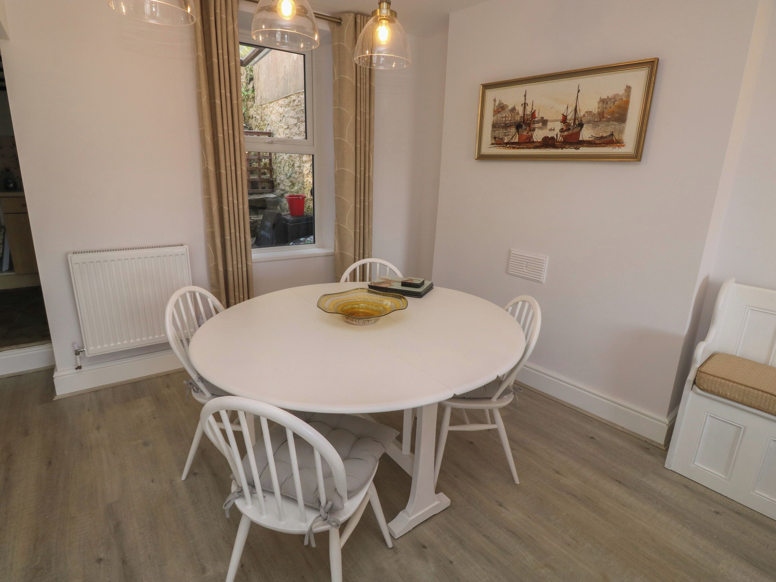 A dining area with a round table, white chairs, a radiator, a window with curtains, and a framed picture on the wall.