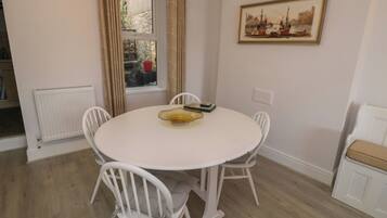 A dining area with a round table, white chairs, a radiator, a window with curtains, and a framed picture on the wall.