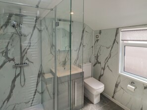 Ein modernes Badezimmer mit einer gläsernen Duschkabine, einer marmorierten Wand, einer weißen Toilette und einem Fenster mit einem gemusterten Vorhang.