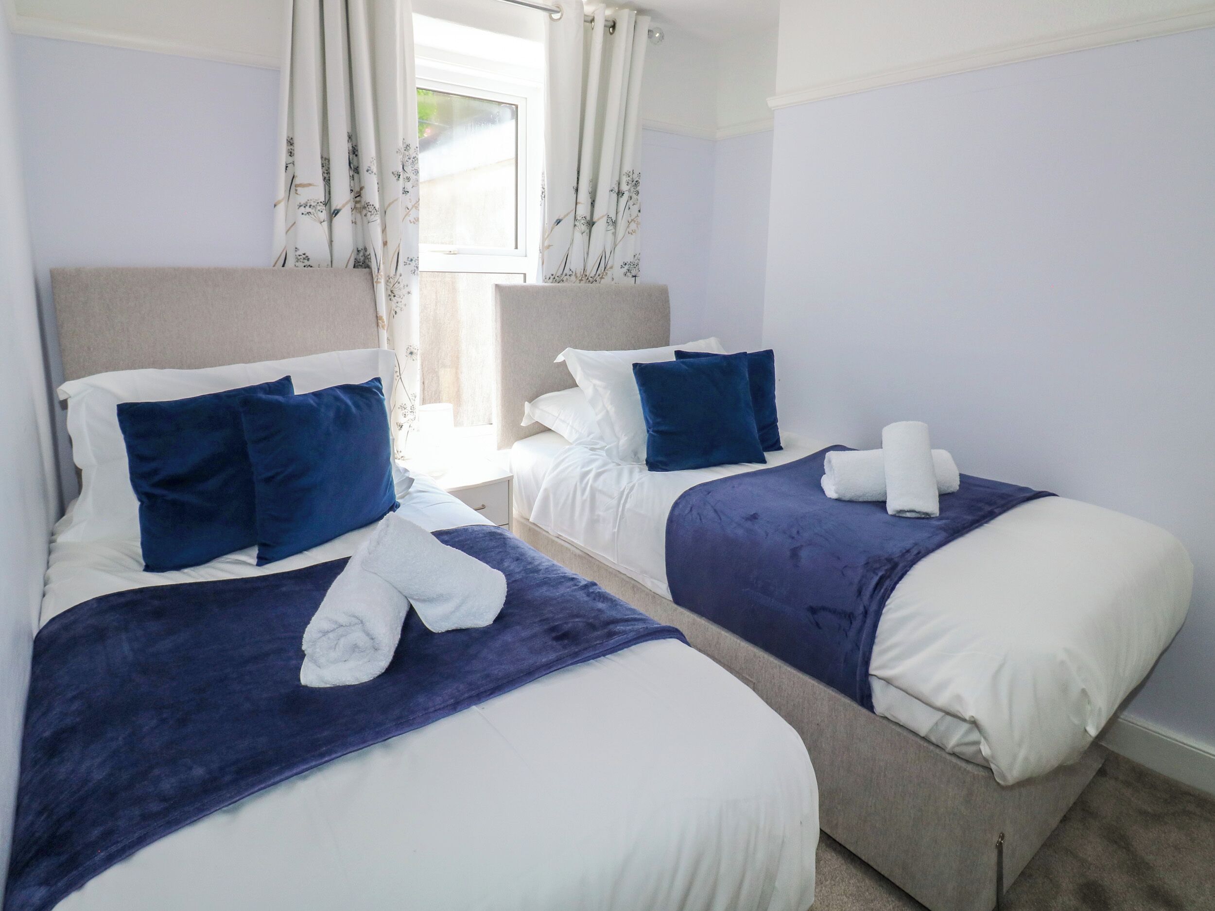 Een hotelkamer met twee bedden, elk voorzien van wit- en blauwe beddengoed, en een raam met bloemen gordijnen.