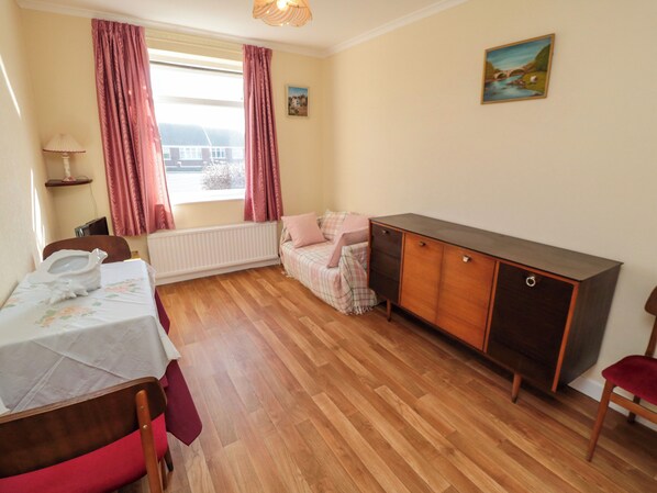 A room with a wooden floor, a bed with a white cover, a wooden cabinet, a chair, a radiator, and a window with curtains. - Sycamore (Filey)