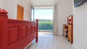Ένα υπνοδωμάτιο με ένα κόκκινο κρεβάτι, μια ξύλινη συρτ αριέρα, μια τηλεόραση τοποθετημένη στον τοίχο και ένα μεγάλο παράθυρο με κουρτίνες.