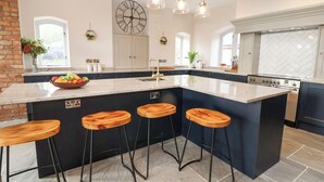 A modern kitchen with a central island, bar stools, pendant lighting, and a large clock on the wall. - Tanat House (Oswestry)