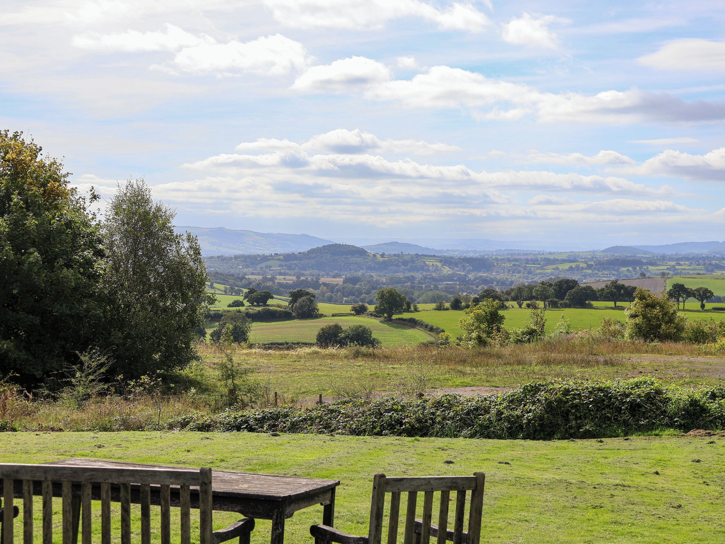 Paisaje rural con un banco y una mesa de madera al frente, un seto y una vista distante de colinas ondulantes y un cielo nublado.