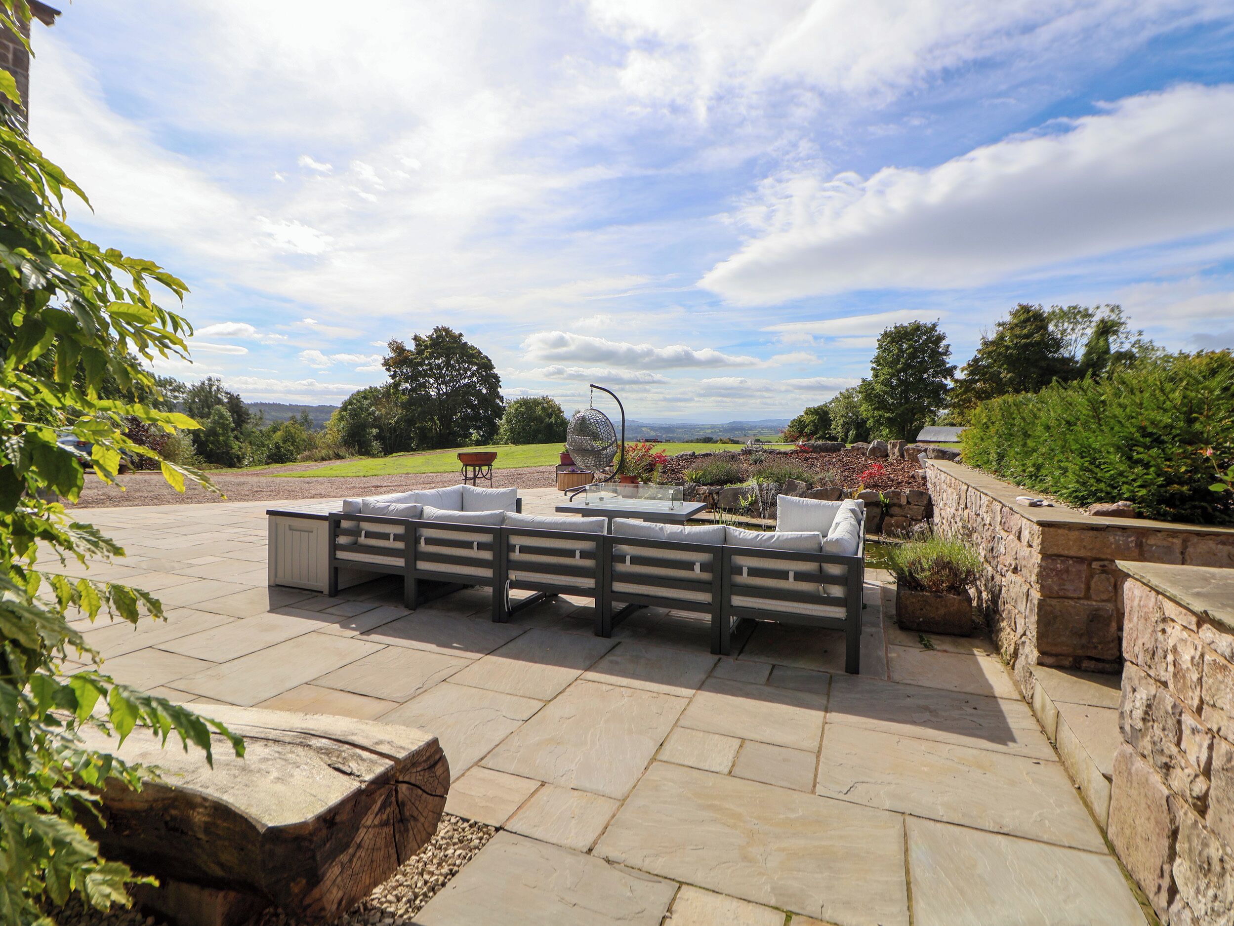 An outdoor patio with a stone table and seating, surrounded by a stone wall and greenery.