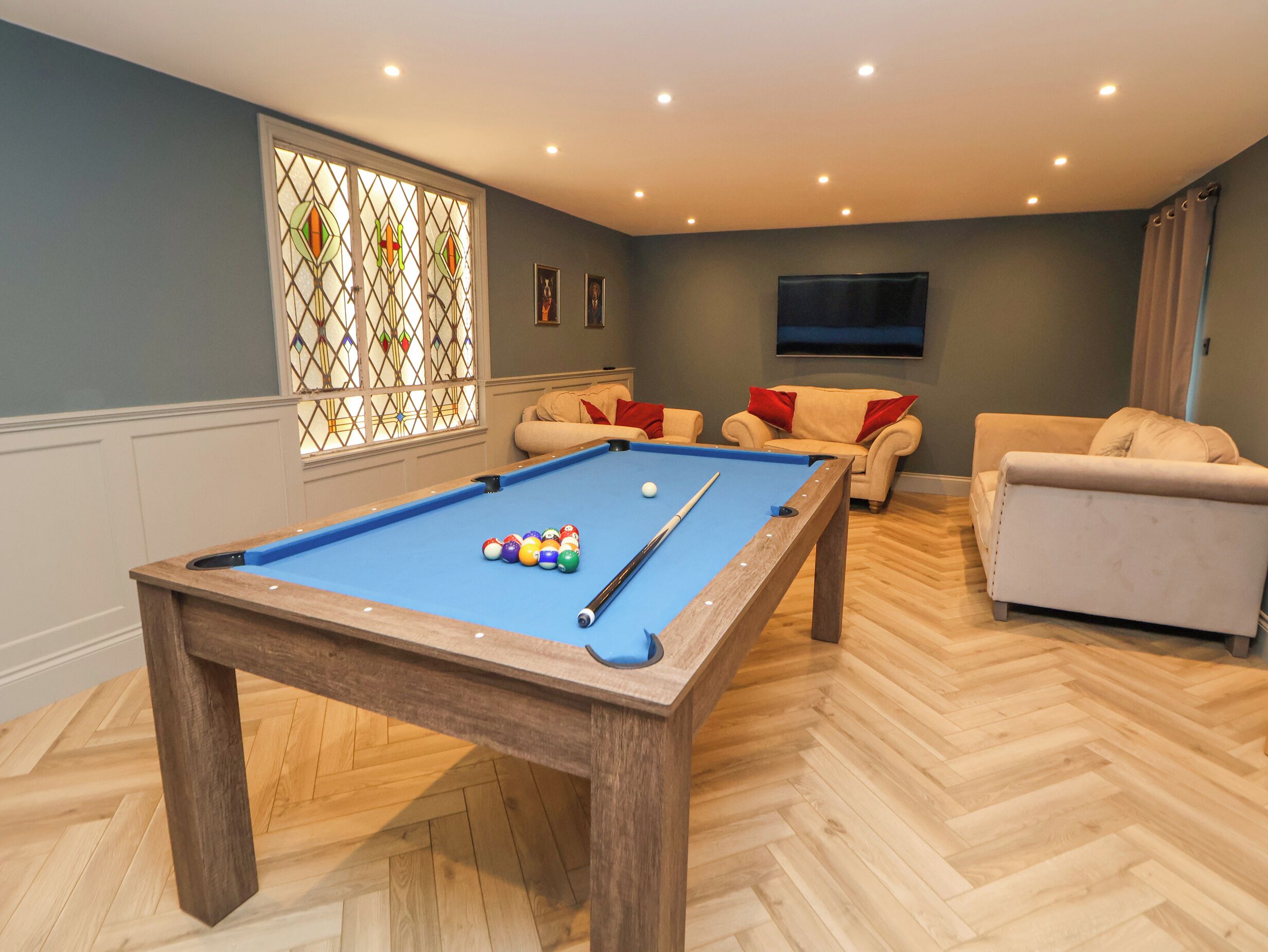 Une table de billard avec des billes, un canapé et un téléviseur dans une pièce dotée d’un plancher en bois et d’une fenêtre avec des vitraux.