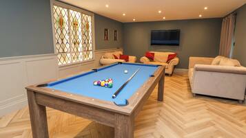 Une table de billard avec des billes, un canapé et un téléviseur dans une pièce dotée d’un plancher en bois et d’une fenêtre avec des vitraux.