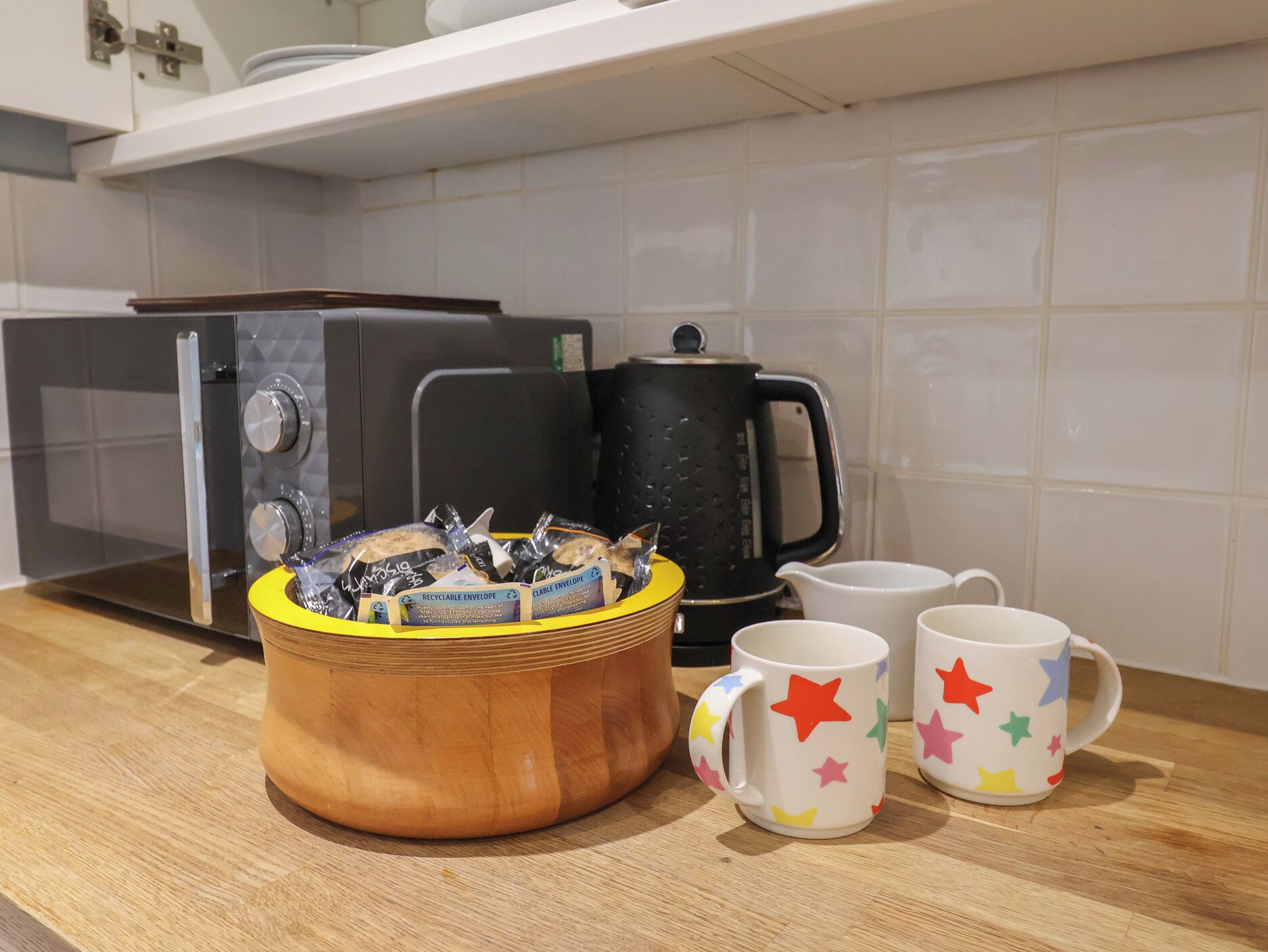 Una encimera de cocina con microondas, hervidor de agua, un cuenco de madera con alimentos y tazas con estampado de estrellas.