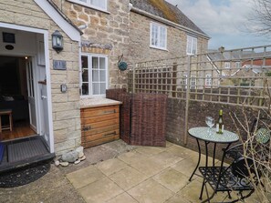 A stone house with a wooden door, a small outdoor seating area with a table and chairs, and a wicker basket. - Old Ropemakers Cottage (Bridport)