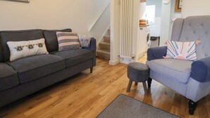 A living room with a grey sofa, a grey armchair, and a small ottoman. There is a staircase leading to another room, and a radiator on the wall. - Old Ropemakers Cottage (Bridport)