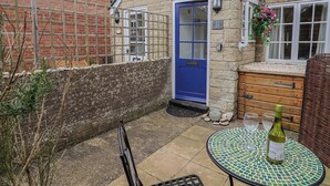 A small stone house with a blue door, a wooden bench, a table with a bottle, and a wicker chair. - Old Ropemakers Cottage (Bridport)