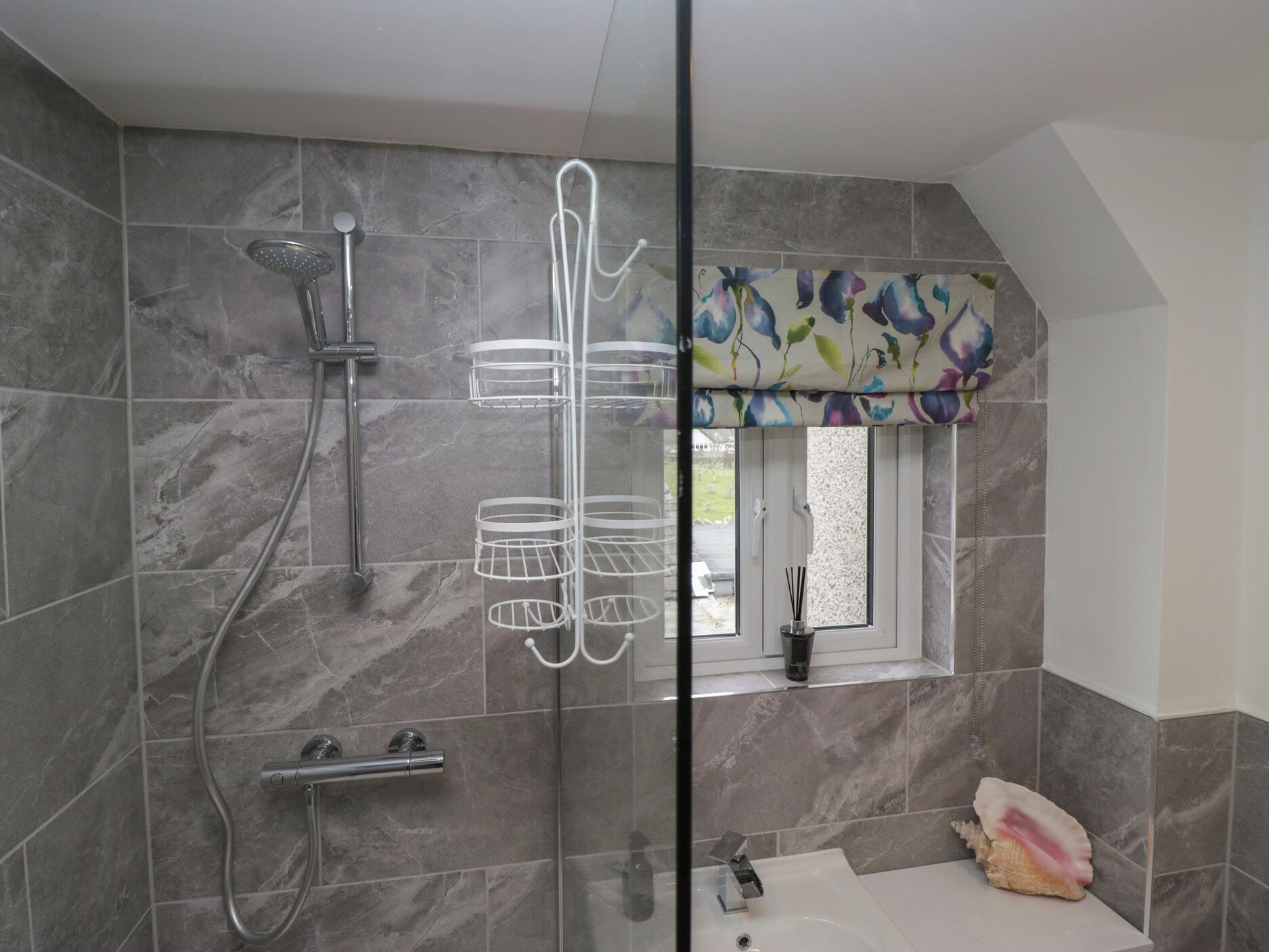 A bathroom with a shower, a sink, and a window with a floral curtain.