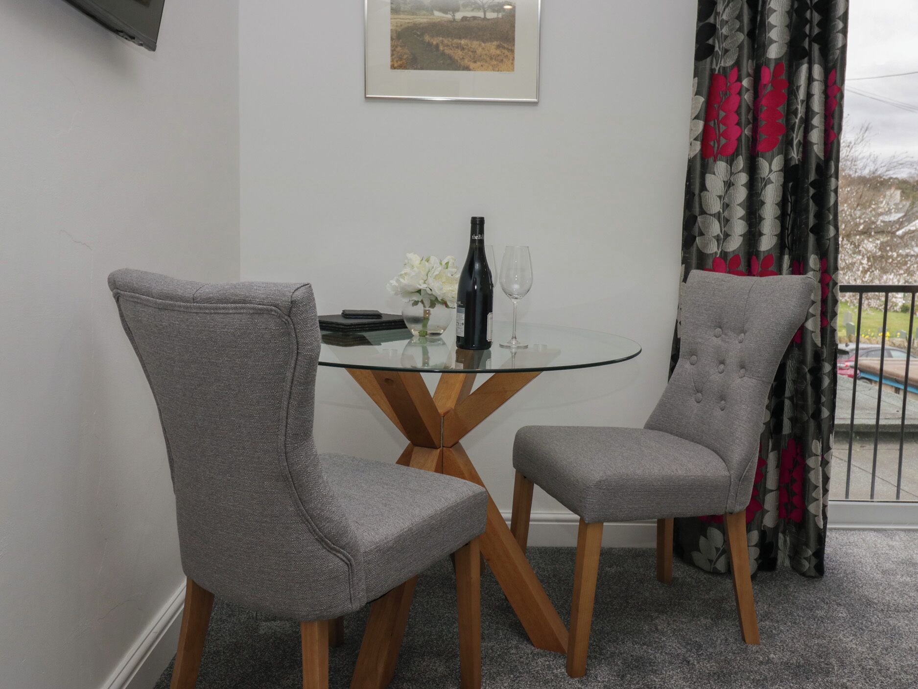 A dining area with two grey upholstered chairs, a glass table, a wine bottle, and a glass.