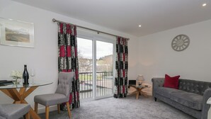 A living room with a grey sofa, a grey armchair, a glass coffee table, and a small side table with a lamp. There is a large window with curtains, a framed picture on the wall, and a clock. - Yewdale View Apartment (Coniston)