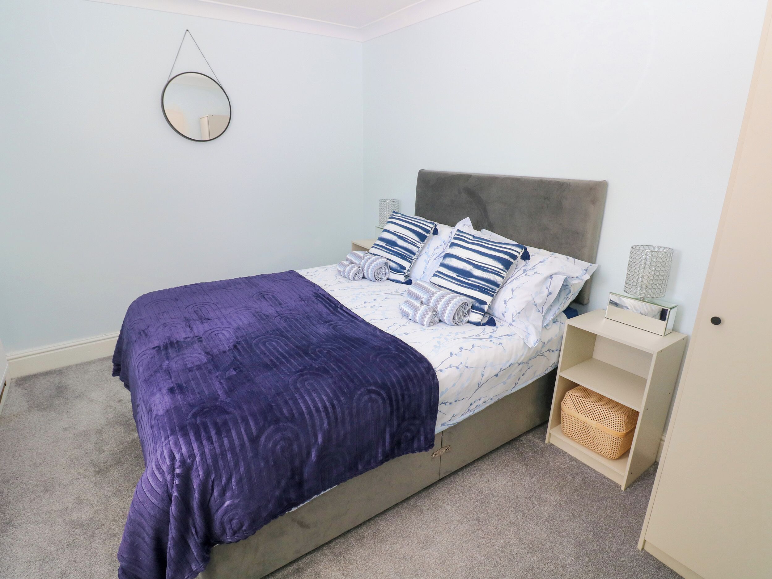 A neatly made bed with a purple blanket, a grey headboard, and striped pillows. There is a bedside table with a lamp and a basket.