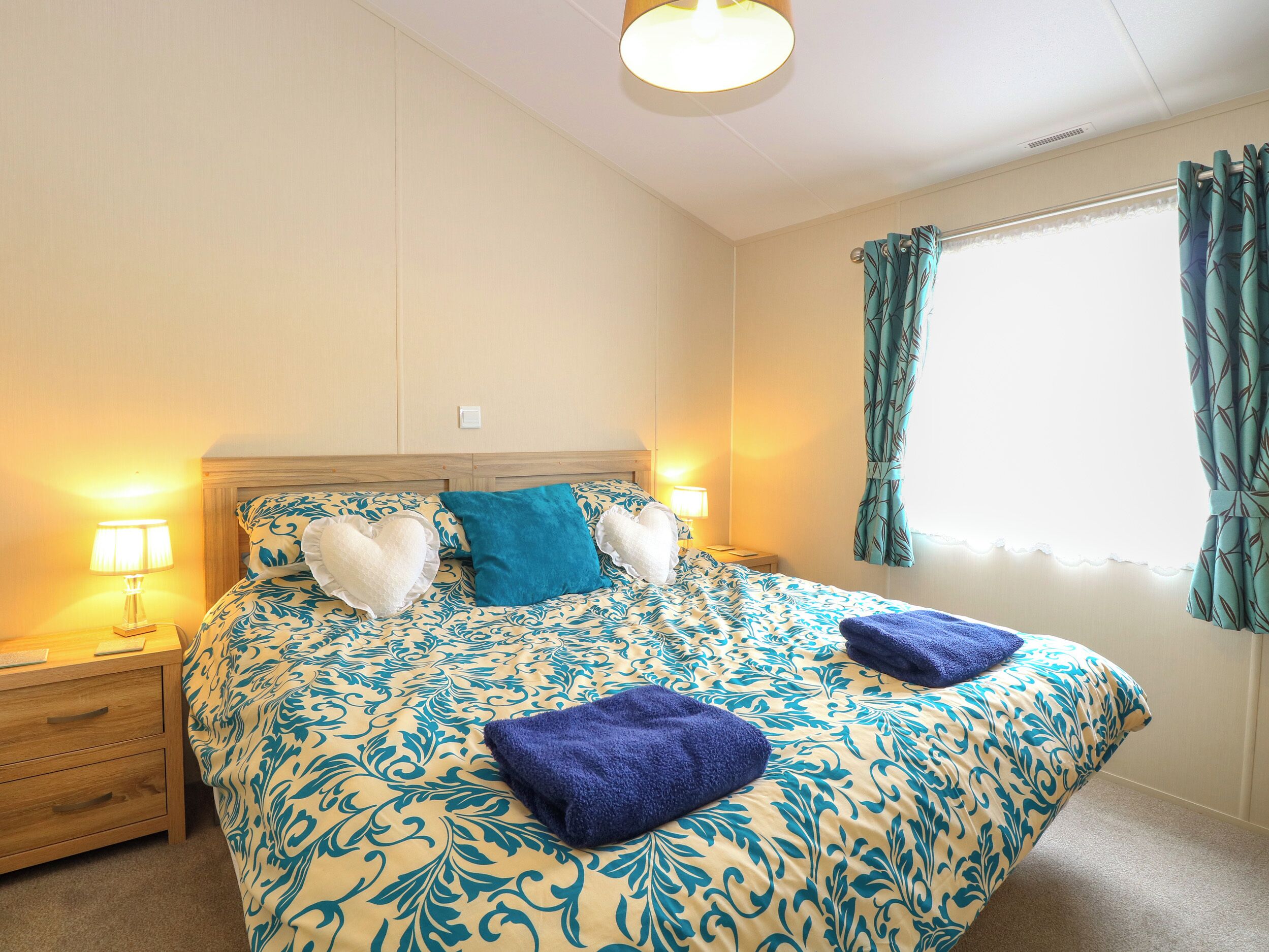 A hotel room with a large bed, two bedside tables, a nightstand, a window with curtains, and a ceiling light.