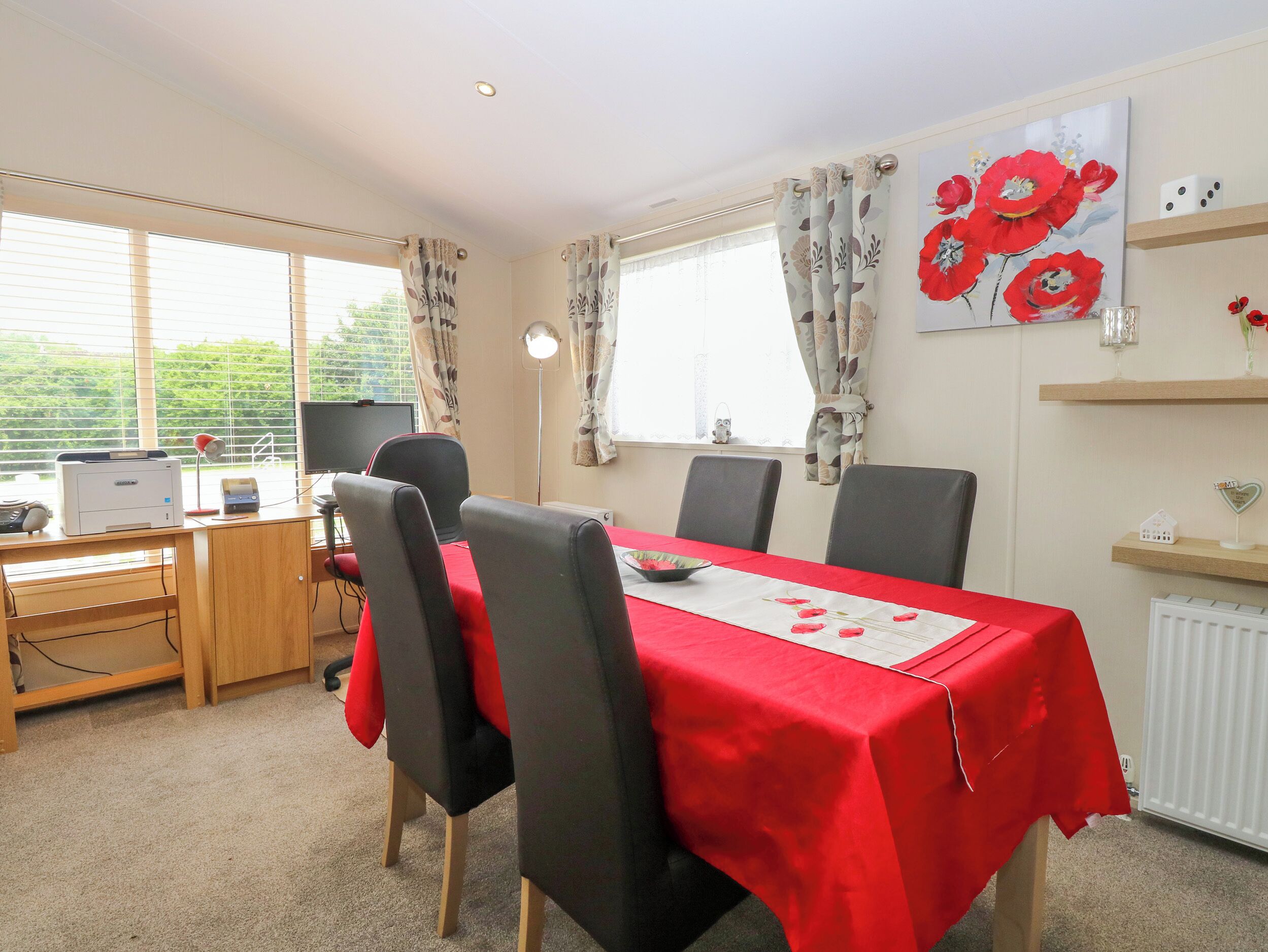 A dining area with a red tablecloth, four chairs, a computer desk with a printer, and a large window with curtains.