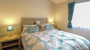 A bedroom with a wooden headboard, a bed with a floral-patterned duvet, two bedside lamps, a nightstand, and a window with curtains. - 21 Norman Drive Lodge (Great Yarmouth)
