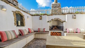 A patio area with stone walls, a white picket fence, a stone chimney, and a seating bench with striped cushions. - Logan Barn (Camelford)