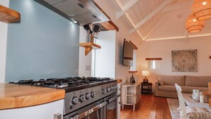 A kitchen with a stove, oven, and wooden countertop; a dining area with a table and chairs; a sofa; a TV; and a patterned wall hanging. - Logan Barn (Camelford)