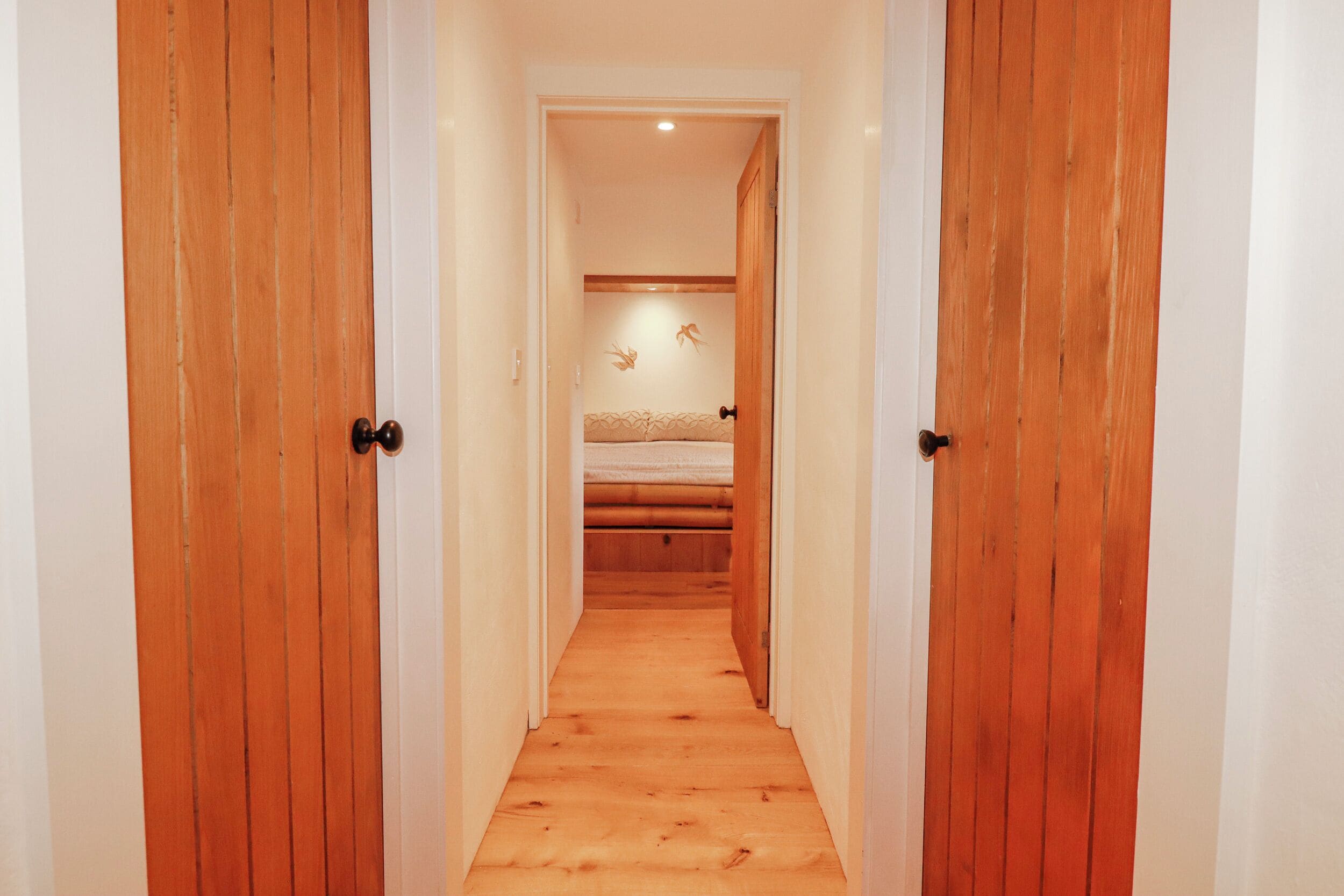 A narrow hallway with wooden doors leading to a bedroom with a bed and a wall-mounted decoration.