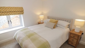 Une chambre à coucher avec un lit, deux tables de chevet avec des lampes, une fenêtre avec un rideau à motifs et une vue sur un bâtiment en briques à l’extérieur.