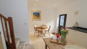 Un coin repas avec une table en bois, des chaises et un vase de fleurs. Un escalier se trouve à gauche, et une cuisine avec une fenêtre et une horloge au mur.