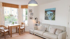 Een gezellige woonkamer met een witte bank, een houten eettafel met stoelen, een hanglamp en een schilderij van een brug.