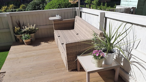 Een houten terras met een rieten bank en een kleine tafel, omringd door potplanten.