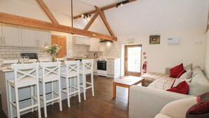 A modern kitchen with white cabinets, a dining area with white chairs, and a living room with a sofa and red cushions. - Millstone Cottages - Breidden View (Shrewsbury)