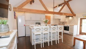 A modern kitchen with white cabinets, wooden beams, a central island with chairs, and a dining table. - Millstone Cottages - Breidden View (Shrewsbury)