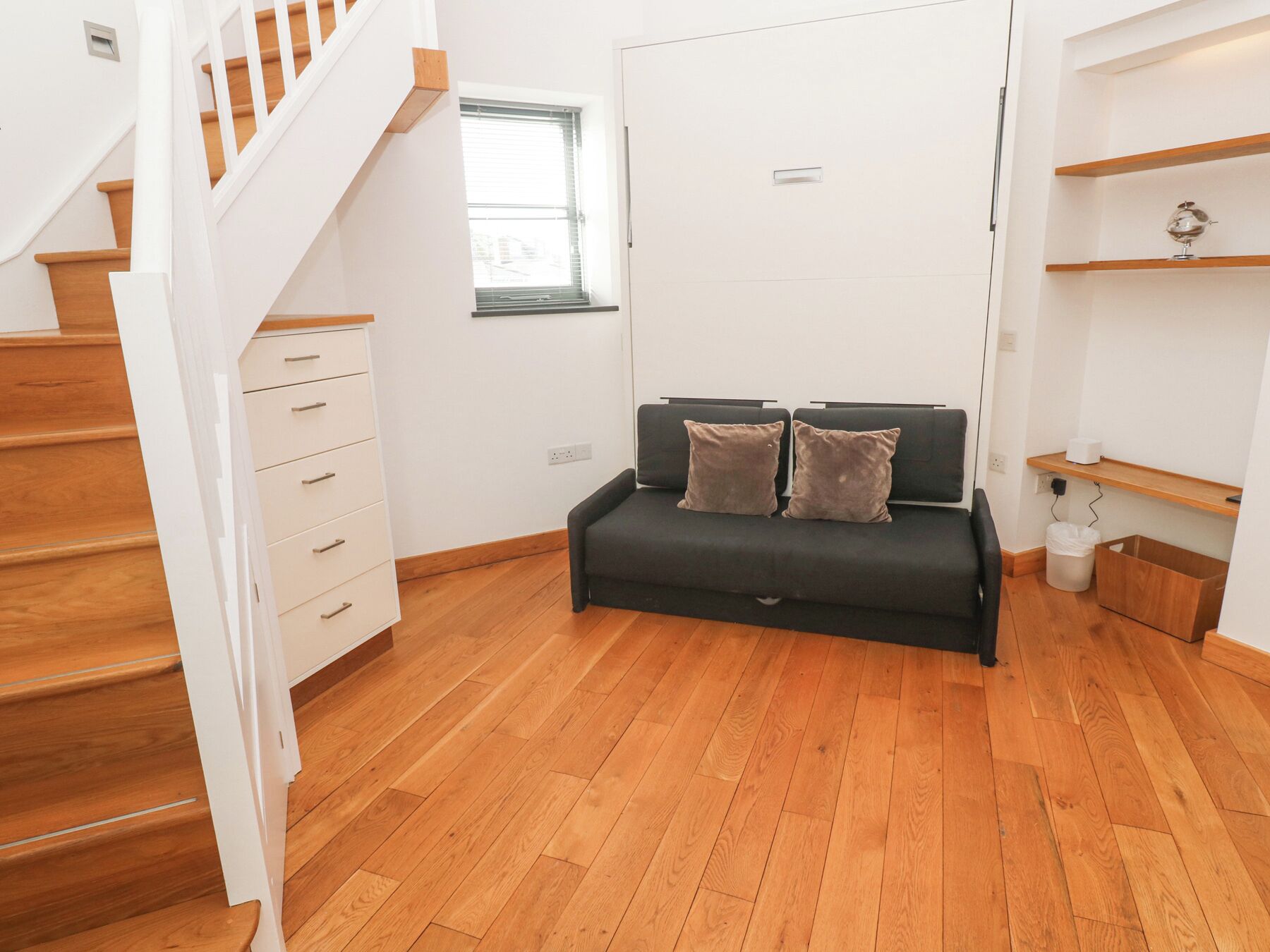 A modern living room with a black sofa, wooden flooring, white walls, a staircase, and a window.