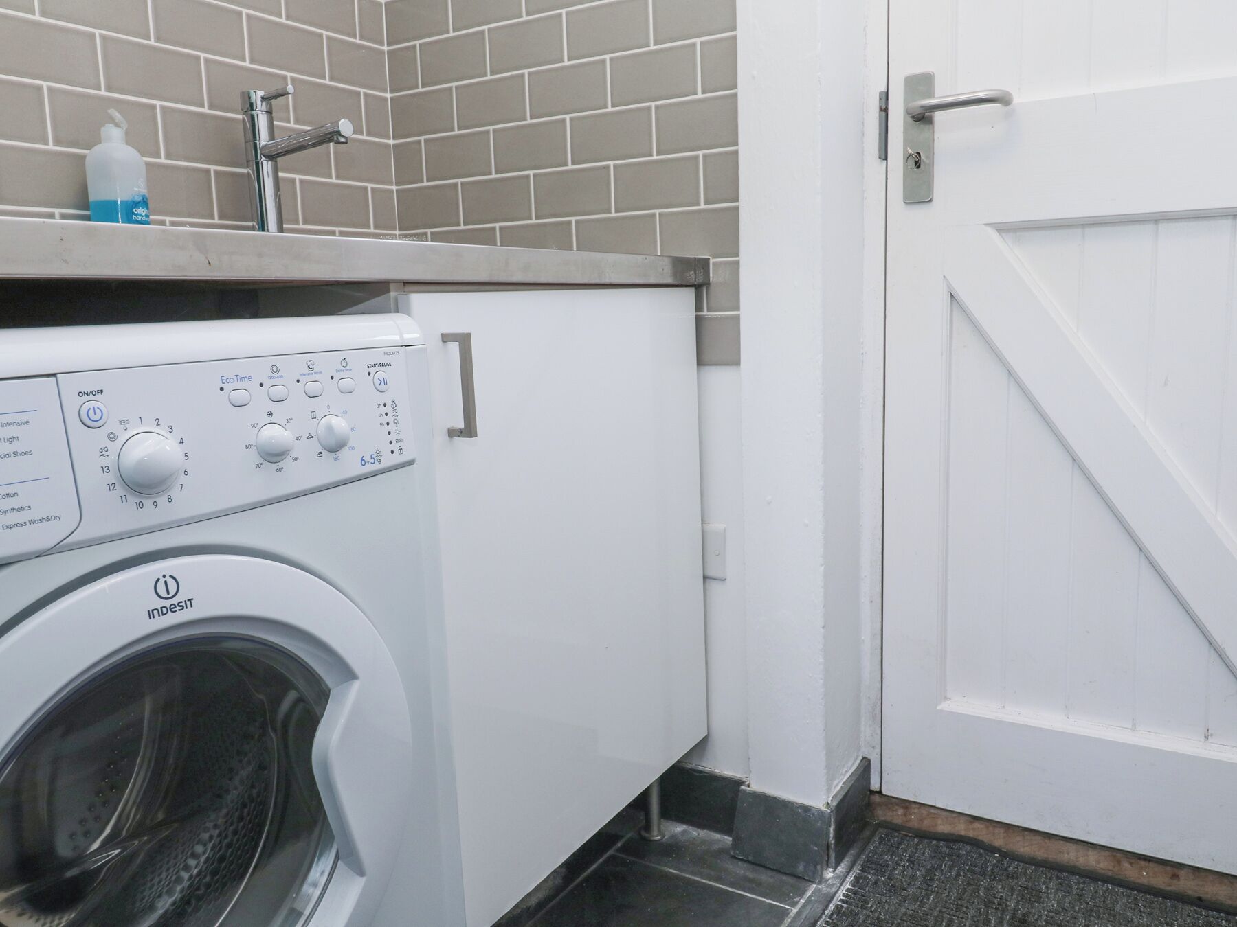 A front-loading washing machine with control panel, a sink with a bottle of soap, and a white door.