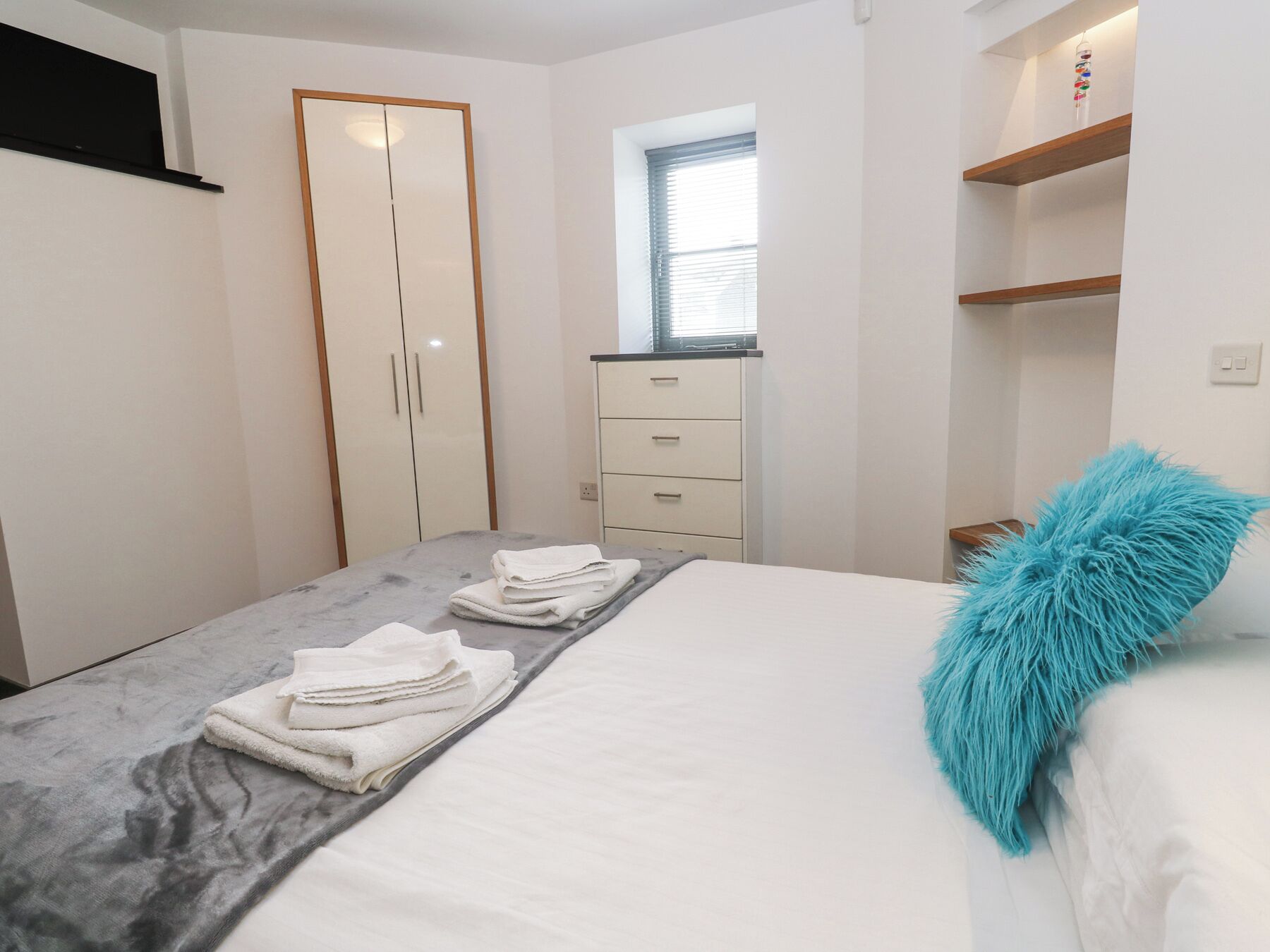 A neatly made bed with white linens and a blue accent pillow, a grey headboard, a wardrobe with a mirror, and a small window with blinds.