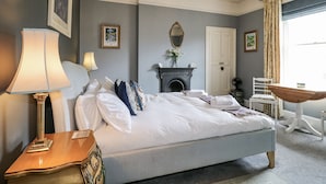 Een opgemaakt bed met witte lakens, een bedlamp, een klein houten nachtkastje, een openhaard, een spiegel en een raam met gordijnen.