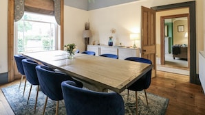 Een eetkamer met een houten tafel en blauwe fauteuils, een kroonluchter en uitzicht door een raam.