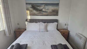 A neatly made bed with white linens, two dark towels, a painting of a boat on the wall, and a radiator on the wall. - Camwy (Llanfairpwllgwyngyll)