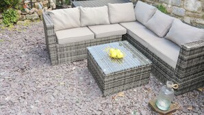 A wicker sectional sofa with cushions, a glass coffee table, and a stone wall in the background. - Camwy (Llanfairpwllgwyngyll)