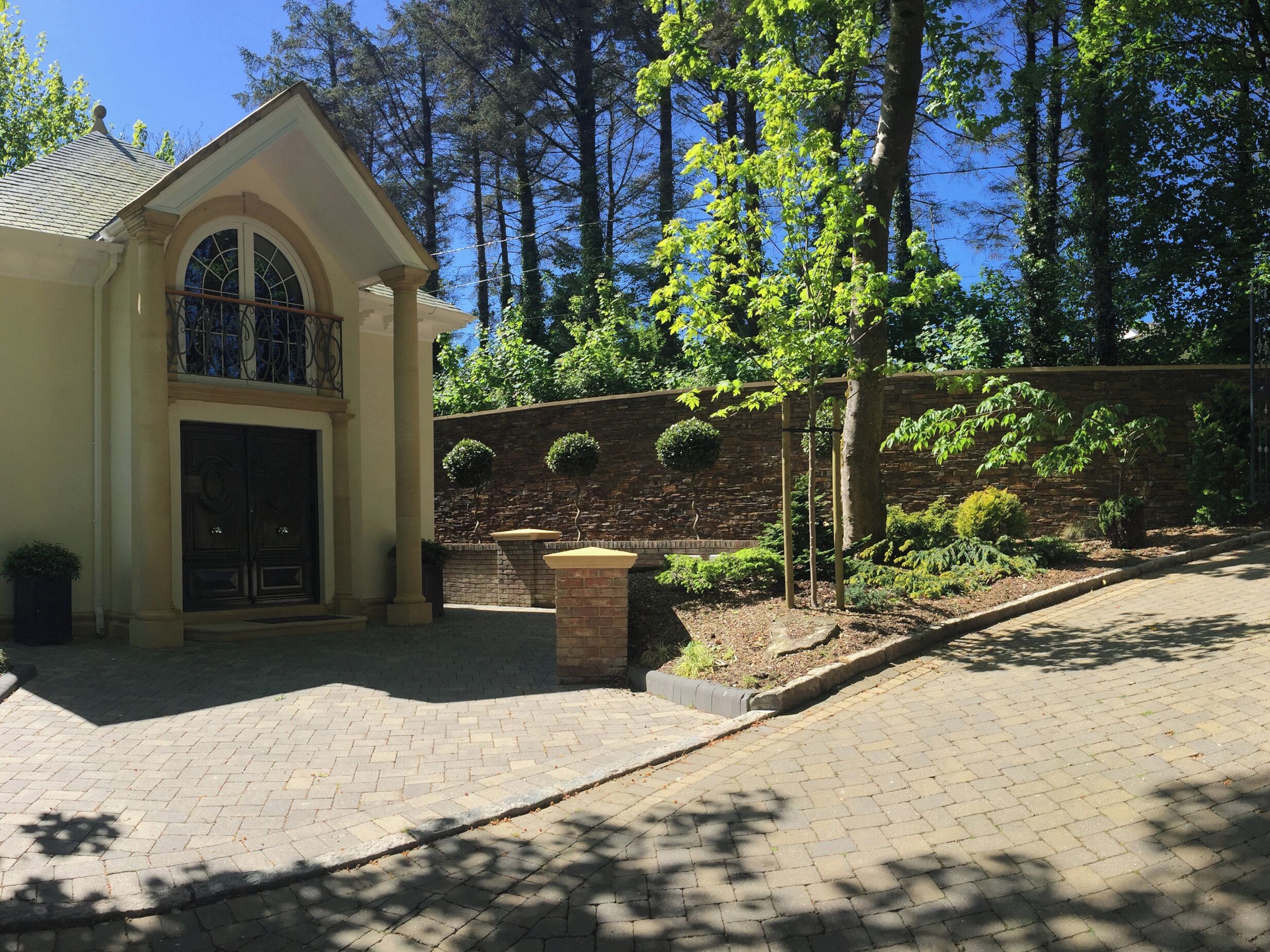A building with a stone facade, arched entrance, and a small garden with a tree and shrubs.