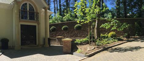 A building with a stone facade, arched entrance, and a small garden with a tree and shrubs.