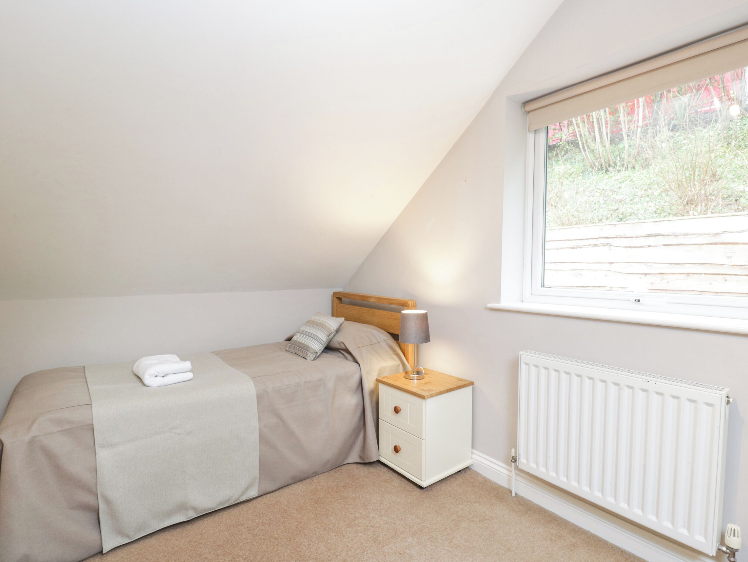 A bedroom with a bed, a nightstand, a lamp, a window, and a radiator.
