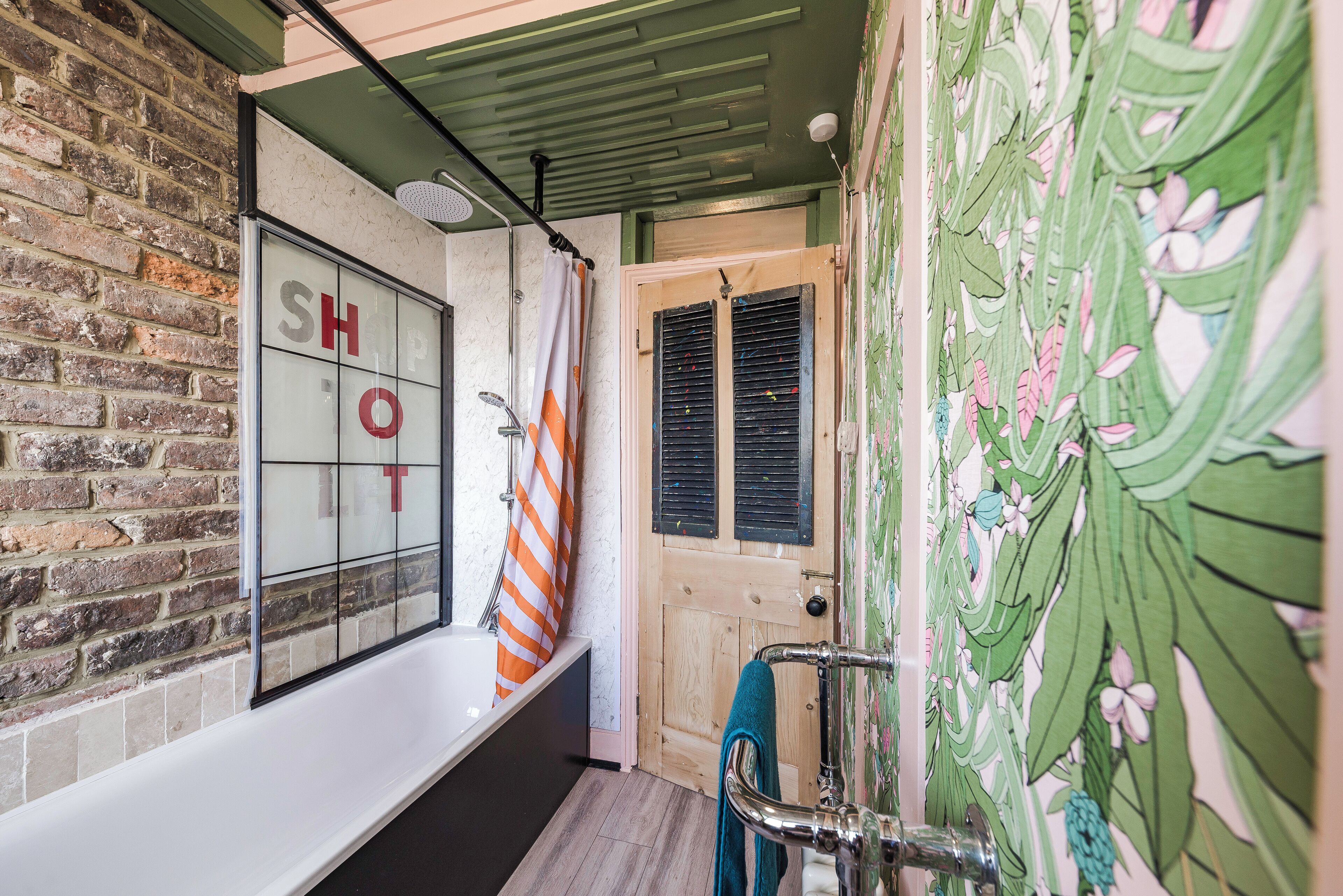 A bathroom with a brick wall, a window with 'SHOP' written on it, a shower with an orange and white striped towel, and a wooden door with shutters.